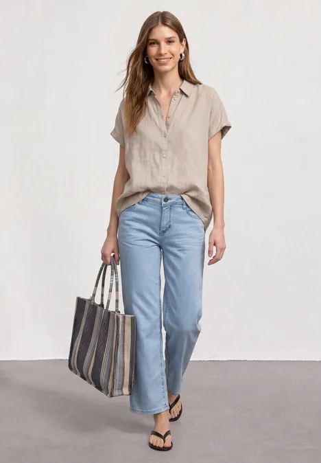 Vrouw in beige blouse en jeans, met zwarte slippers en gestreepte tas, loopt ontspannen.