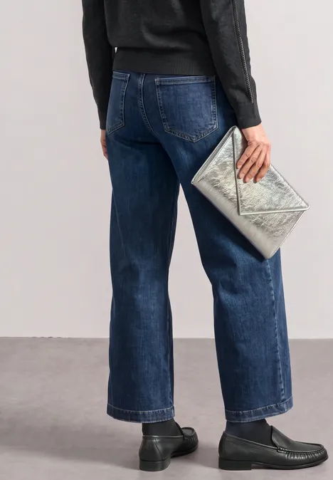 Person in weiter geschnittenen Jeans und schwarzem Oberteil hält silberne Clutch.