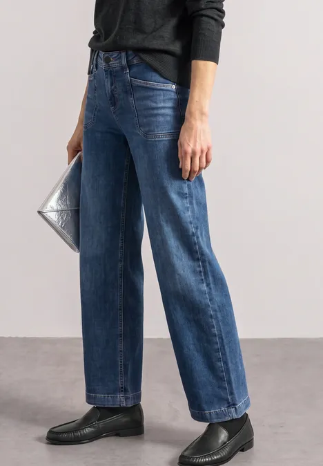 Frau trägt dunkelblaue, weite Jeans, schwarze Loafer und hält eine silberne Clutch.