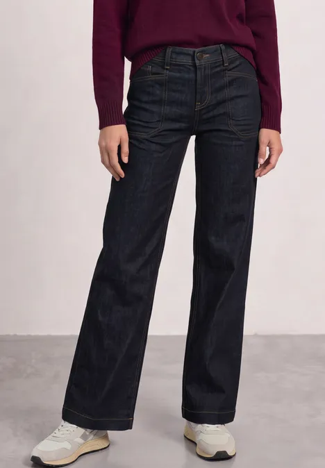 Persoon draagt wijde donkerblauwe jeans en witte sneakers tegen een neutrale achtergrond.