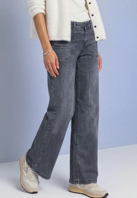 Mid Waist Wide Leg Jeans im Loose Fit Steel Grey Washed Unterkörper einer Person in grauen, weiten Jeans und weißen Sportschuhen vor blauem Hintergrund.