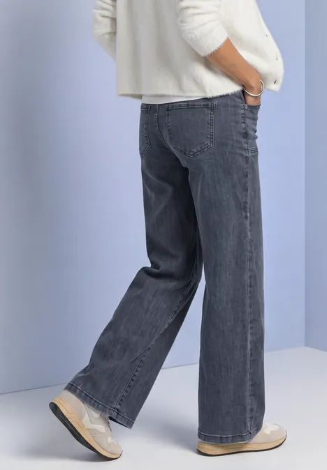 Mid Waist Wide Leg Jeans im Loose Fit Steel Grey Washed Person steht in weitem grauen Jeans mit Händen in den Taschen, weiße Schuhe, vor hellem Hintergrund.