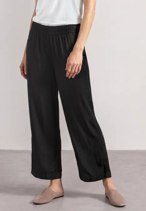 Schwarze, weite Hose mit Gummibund und grauen Slippern vor neutralem Hintergrund.