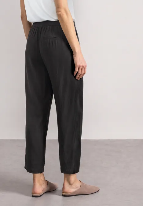 Ein Modell von hinten in schwarzen, lässigen, hoch geschnittenen Hosen mit elastischem Bund und beigen Slippern.