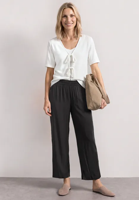 Frau trägt weißes T-Shirt, schwarze weite Hose, beige Schuhe, hält Papiertüte.