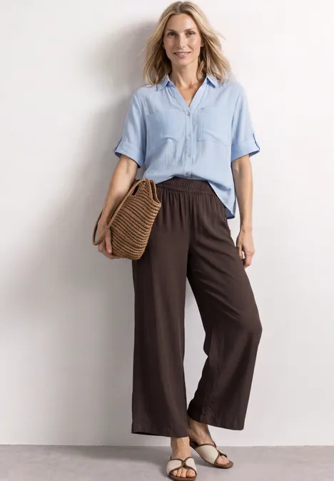 Frau in hellblauer Bluse und weiten braunen Hosen mit Basttasche und Sandalen vor neutralem Hintergrund.