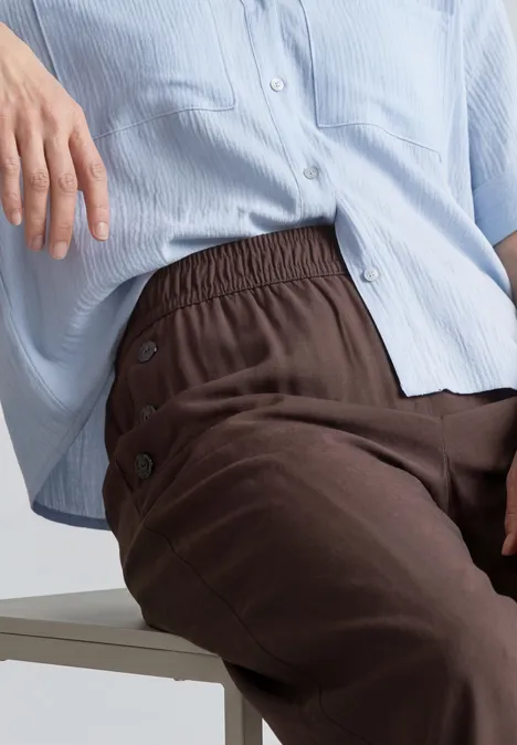 Person sitzt, trägt ein lockeres hellblaues Hemd und eine dunkelbraune Hose mit seitlichen Knöpfen.