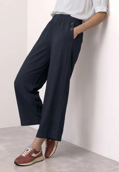 High Waist Wide Leg Hose im Loose Fit shadowed navy Person lehnt an weißer Wand, trägt schwarze, weite Hose mit Taschen und rote Sneaker.