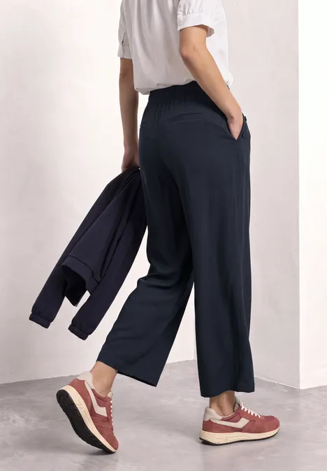 High Waist Wide Leg Hose im Loose Fit shadowed navy Person von hinten in legerer, weiter dunkler Hose, weißem T-Shirt, rosa Sneakern und Jacke in der Hand.