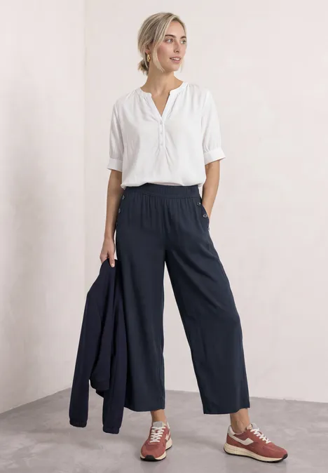 High Waist Wide Leg Hose im Loose Fit shadowed navy Frau in weißer Bluse und dunkelblauen Culottes hält eine Jacke, trägt braune Schuhe.
