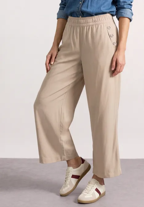 Frau trägt eine beige Culotte mit elastischem Bund und seitlichen Knöpfen, kombiniert mit weißen Sneakern mit Streifen.