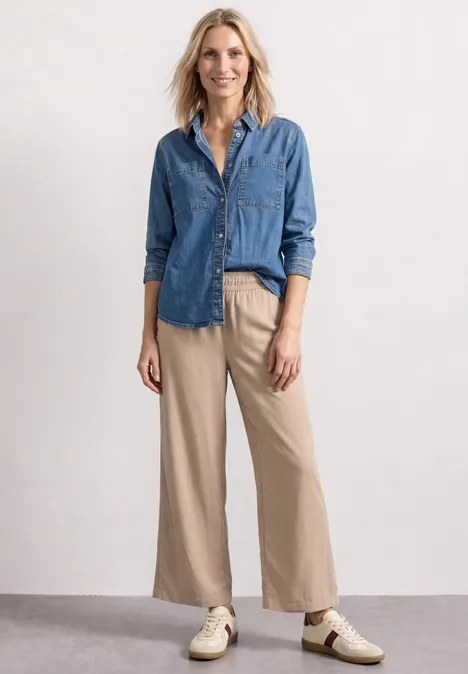 Eine Frau in einem blauen Jeanshemd, beigefarbenen, weiten Hosen und Sneakers steht lässig.