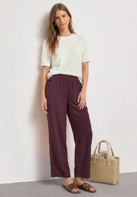 Dame trägt weißes T-Shirt und weinrote Hose, Sandalen, neben ihr eine Basttasche.
