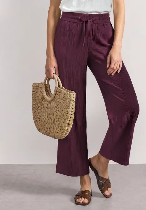 Frau trägt lila weite Hose mit weißem Top, braunen Sandalen und geflochtenen Basttasche.