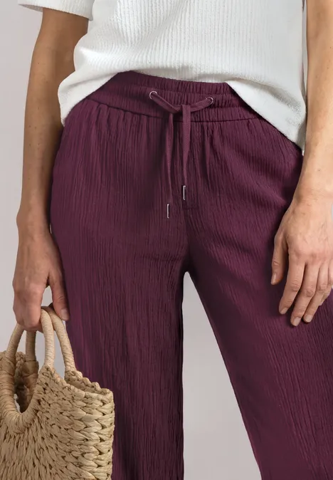 Weite, gerippte bordeauxrote Stoffhose mit Tunnelzug, weißer Top und Korbtasche.