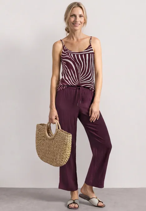 Frau trägt gestreiftes Top, breite lila Hose, Sandalen und eine Strohtasche.