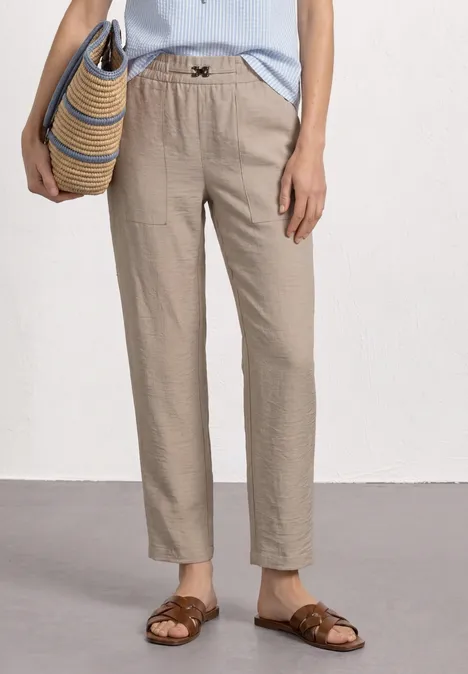 Frau mit beiger Leinenhose, hellblauem Top, braunen Sandalen und einer geflochtenen Strohtasche.
