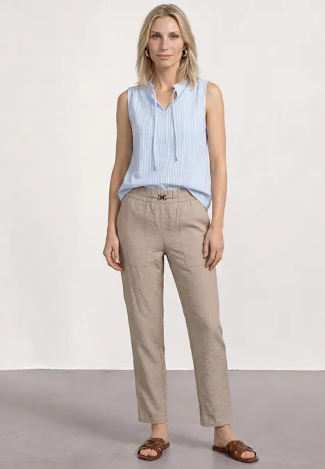 Frau trägt hellblaues ärmelloses Top, beige Hose und braune Sandalen vor schlichtem Hintergrund.