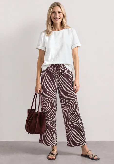 Wide Leg Hose mit Print und Gallonstreifen rustic rouge Eine Person mit weißem T-Shirt, zebraähnlicher, weiter Hose und Sandalen, trägt eine braune Tasche.