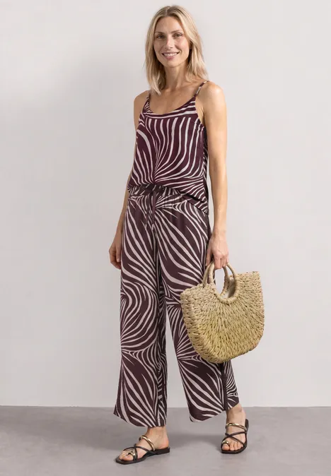 Wide Leg Hose mit Print und Gallonstreifen rustic rouge Frau in gestreiftem, zebraähnlichem Top und Hose, trägt Strohtasche und schwarze Sandalen vor neutralem Hintergrund.