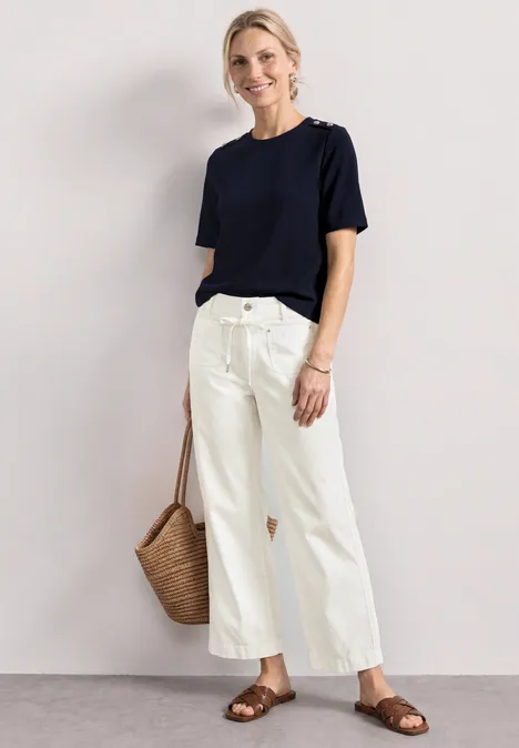 Person in schlichtem dunkelblauen T-Shirt, weiter weißer Hose, braunen Sandalen und geflochtener Strohtasche.