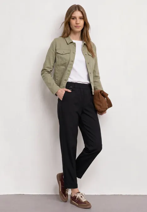 Frau vor weißer Wand, grüne Jacke, weißes Shirt, schwarze Hose, braune Schuhe und Tasche.