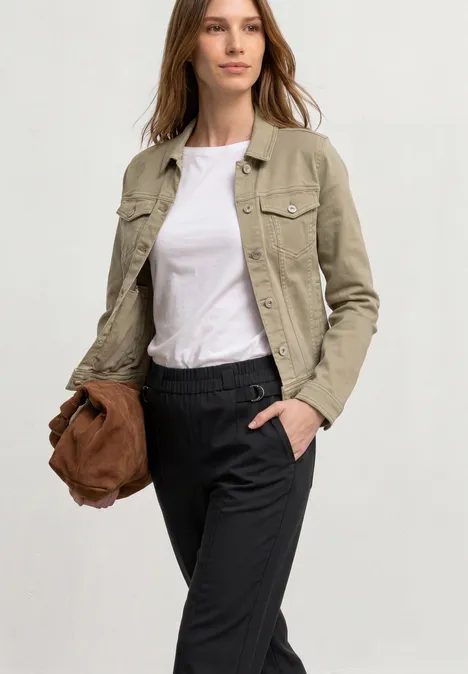 Frau in hellbeiger Jacke, weißem T-Shirt und schwarzen Hosen hält braune Tasche, Hand in der Tasche.