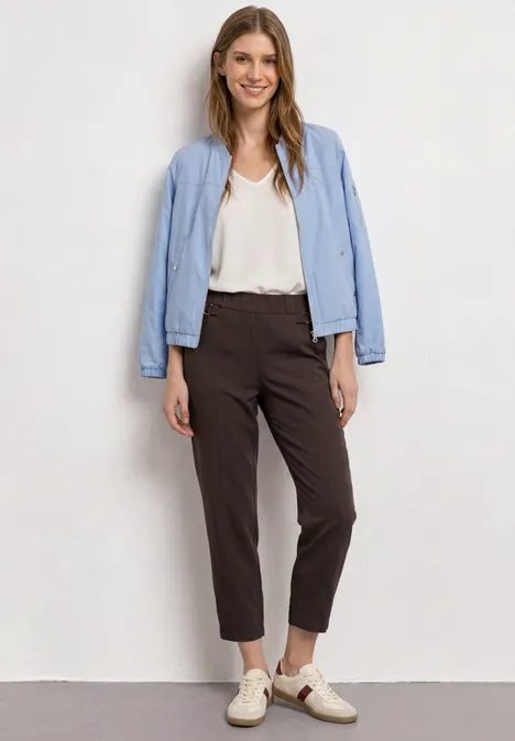 Vollkörperfoto einer Frau in weißer Bluse, hellblauer Jacke, braunen Chinos und hellen Slippern vor weißer Wand