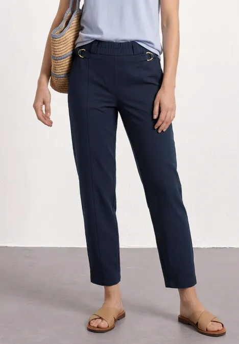 Dunkelblaue, gerade geschnittene Hose, flache Sandalen und geflochtene Tasche an der Hüfte.