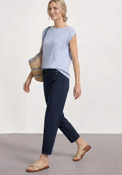 Frau im hellblauen Top, dunkelblauer Hose, flachen Sandalen und Strohtasche.