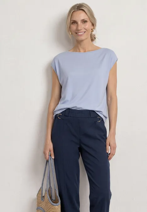 Frau vor weißer Wand in hellblauem Shirt, dunkelblauen Hosen und gestreifter Tasche.