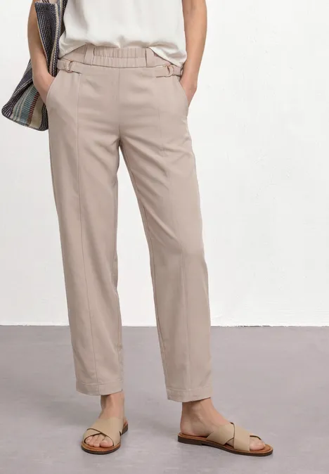 Beine einer Frau in beigefarbenen Hosen, weißem Top, offenen Sandalen, lässige Plaid-Umhängetasche.