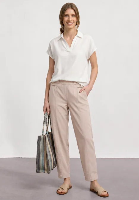 Eine Person trägt ein weißes, kurzärmeliges Hemd, beige Hose, Sandalen und eine gestreifte Tragetasche.