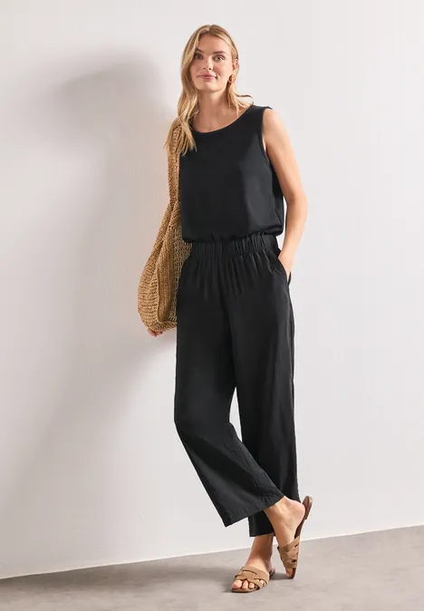 Wide Leg Jumpsuit im Strukturmix Black Frau in lockerem schwarzen Jumpsuit mit Taschen, Strohtasche und Sandalen vor weißer Wand.