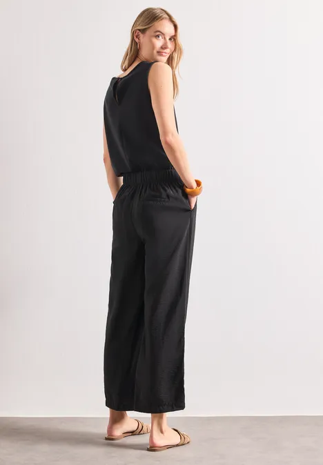 Wide Leg Jumpsuit im Strukturmix Black Frau in schwarzem ärmellosem Jumpsuit mit weitem Bein und Sandalen, seitlich vor neutralem Hintergrund.