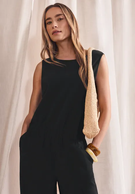 Wide Leg Jumpsuit im Strukturmix Black Frau trägt schwarzen ärmellosen Jumpsuit mit Gummibund und Taschen, hält grob gestrickte Schultertasche.