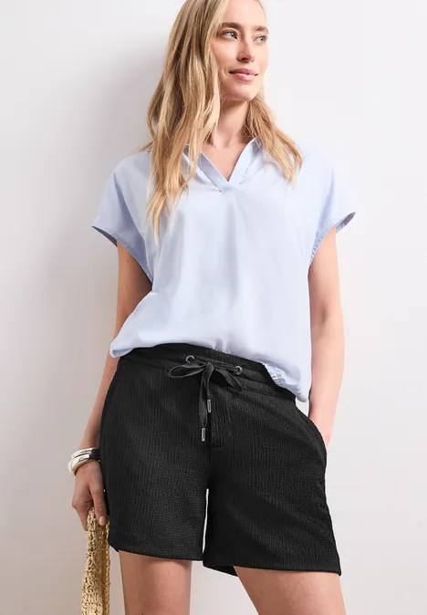 Frau in hellblauer Bluse, schwarzen Shorts und mit Strohtasche.