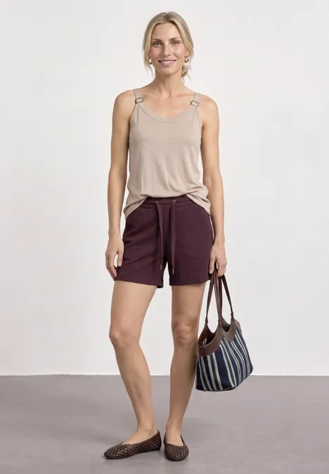 Eine Frau steht vor weißer Wand, trägt beige Top, dunkelrote Shorts, flache Schuhe und hält eine gestreifte Tasche.