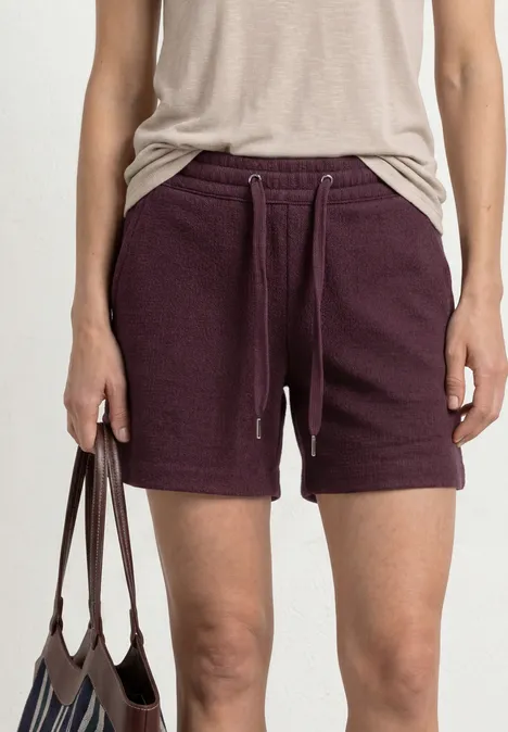 Person trägt beiges Top und dunkelviolette Sweatshorts mit Tunnelzug, hält braune Tasche.