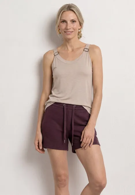 Eine Frau trägt ein hellbeiges Tanktop und dunkelrote Shorts mit Tunnelzug vor hellem Hintergrund.