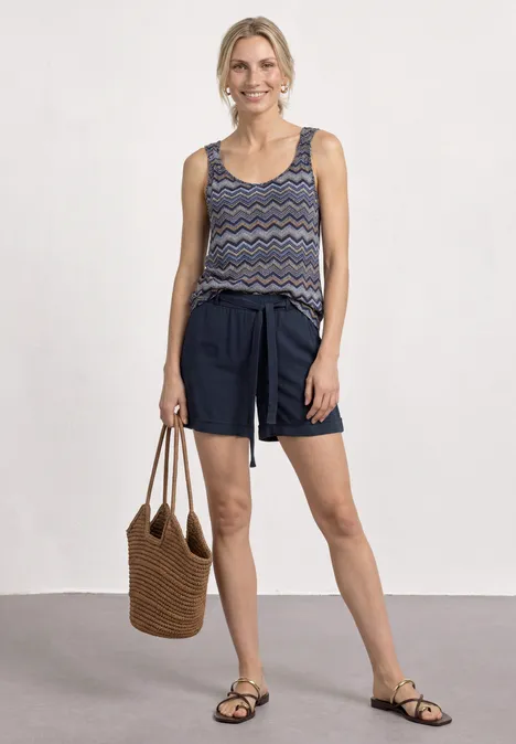Frau in gemustertem Top, dunkelblauen Shorts, Sandalen und geflochtener Tasche.
