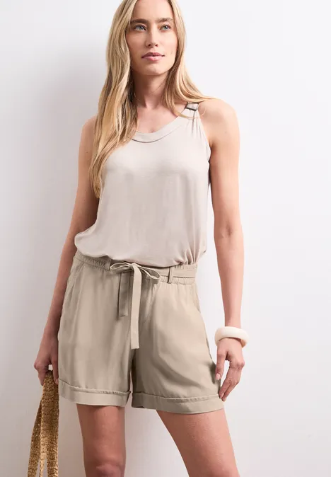 Frau im beigen Tanktop und Shorts mit Strohtasche vor hellem Hintergrund.