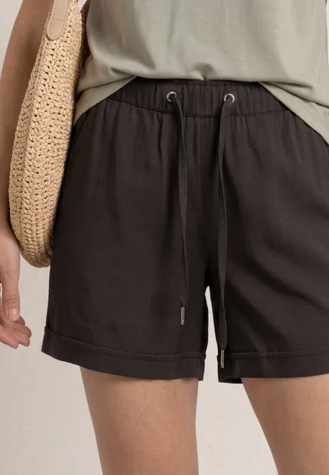 Person in lockeren schwarzen Shorts mit Tunnelzug, grünem Top und geflochtener Tasche aus Bast.