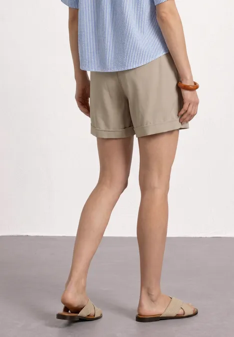 Person in beigen Shorts, blauem Hemd und braunen Sandalen, stehend auf grauem Boden, Rückansicht der Beine.