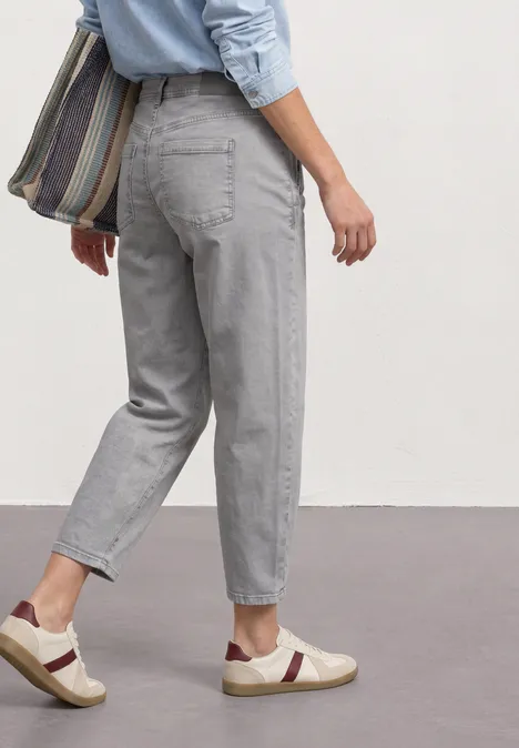 Eine Person trägt hellgraue Jeans, weiße Sneaker mit dunklen Streifen und eine gestreifte Umhängetasche.