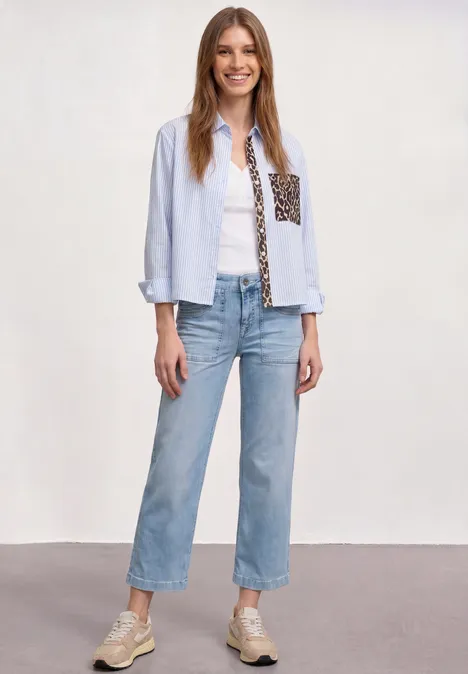 Mid Waist Jeans Culotte im Loose Fit Ultra Bleached Random Wash Dame in hellblauem Hemd mit Muster, weißem Top, geraden blauen Jeans und Sneakers.