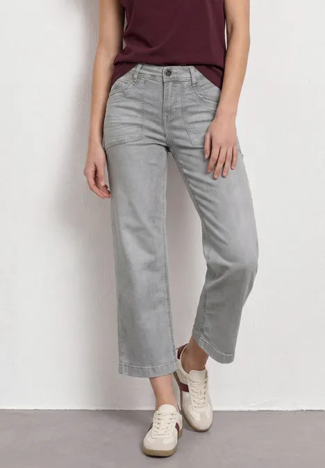 Mid Waist Jeans Culotte im Loose Fit Light Grey Random Frau in grauen, weit geschnittenen Jeans, burgunderrotem Oberteil und weißen Sneakern vor weißem Hintergrund.