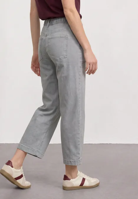 Mid Waist Jeans Culotte im Loose Fit Light Grey Random Frau im Profil trägt hellgraue, hochgeschnittene, weite 7/8-Jeans und weiße Sneaker.
