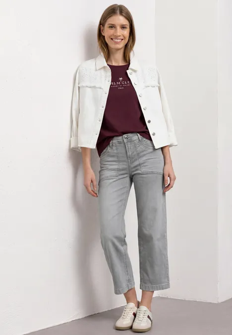 Mid Waist Jeans Culotte im Loose Fit Light Grey Random Frau vor weißer Wand, trägt weiße Jeansjacke, burgundfarbenes Shirt, graue weite Jeans und weiße Sneaker.