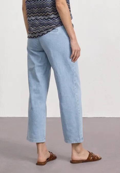 Frau von hinten in hellblauen, weiten Jeans und braunen Sandalen vor weißer Wand.